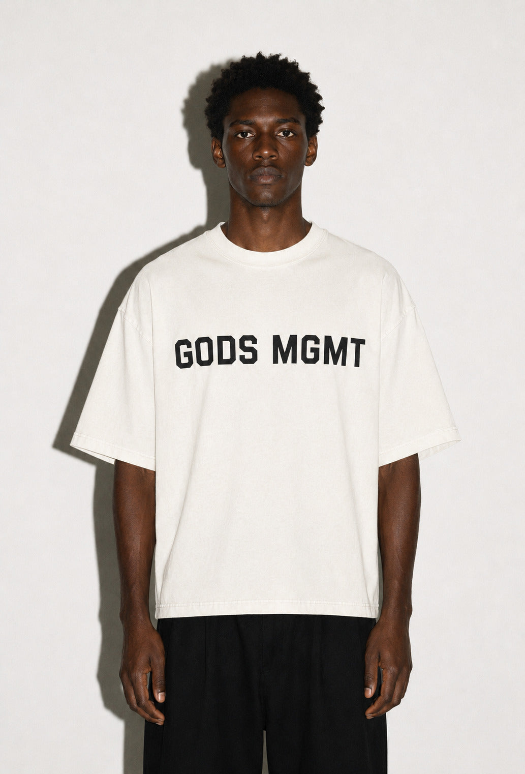 GODS MGMT UNIFORM 001 short sleeve tee in bone, worn against a studio wall